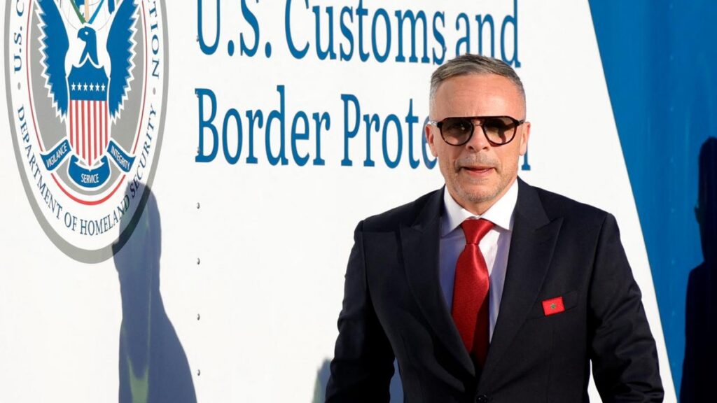 Homme portant une cravate rouge devant un panneau U.S. Customs and Border Protection, avec un pin du Maroc